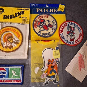 Könnte beinhalten: Eine Sammlung gestickter Aufnäher mit verschiedenen Designs, darunter ein indianischer Häuptling, eine Comicfigur mit herausgestreckter Zunge, Bugs Bunny als Onkel Sam und eine Comicfigur mit Bart. Die Aufnäher befinden sich auf gelber und weißer Verpackung mit dem Text "The Show Offs", "Embroidered Emblems", "What You See Is What You Get", "Great America" und "Wrights Applique Applicazione".