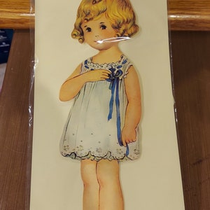 Puede incluir: Un conjunto de muñecas de papel vintage con una niña llamada "Dolly Dimple" con cabello rubio y un vestido azul. El conjunto incluye una muñeca de 23 cm, 5 trajes y 5 sombreros. El embalaje es rojo y blanco con el texto "The Boston Children's Museum".
