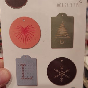 May include: A set of six die-cut metal shapes for crafting. The shapes include a heart, a Christmas tree, a snowflake, and a letter L. The shapes are all in different colors and have a stitched design. The package is labeled "Sizzix Thinlits" and "Josh Griffiths".