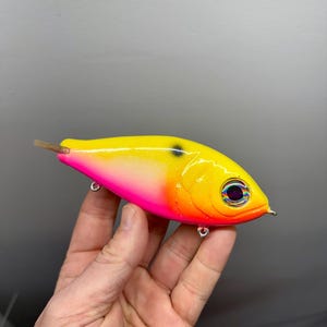 May include: A vibrant fishing lure with a yellow top, transitioning to pink and orange. It features a large, detailed eye and silver hooks. The lure has a small, brown tail and is designed for attracting fish.