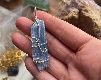Puede incluir: Un colgante de cristal de cianita azul envuelto en alambre plateado. El cristal es largo, delgado y puntiagudo.