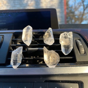 May include: Five clear quartz crystal points are arranged in a car's air vent. The crystals are pointed upwards and are reflecting the sunlight.