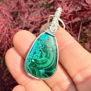 May include: A teardrop-shaped pendant featuring a polished malachite stone with swirling green and teal patterns. The stone is encased in a silver wire wrap, with a silver bail for a necklace chain. The background is a blurred view of red leaves.