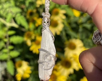 May include: A white selenite crystal pendant wrapped in silver wire. The crystal is long and thin, with a pointed tip. The wire is wrapped around the crystal in a decorative pattern.