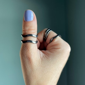 May include: A hand with a light purple painted fingernail and several silver-colored rings. Two rings are on the thumb, with a wavy design. Other rings are on the adjacent fingers.