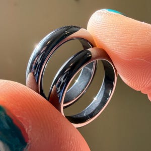 May include: Two black and silver rings are held between fingers. The rings have a shiny, reflective surface. The rings are likely made of metal, and the design is simple and modern. The rings are of different sizes, suggesting they are a set.