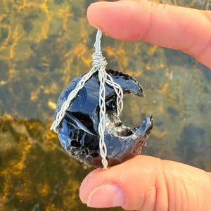 May include: A black obsidian crescent moon pendant with a silver wire wrap. The pendant is held by a finger against a blurred background of water.