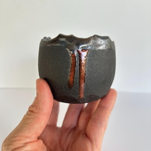 May include: A small, round ceramic cup with a matte black, textured exterior. The rim features a glossy, dark metallic glaze that drips down the side in two distinct, vertical streaks of reddish-brown and orange, creating a contrast in texture.