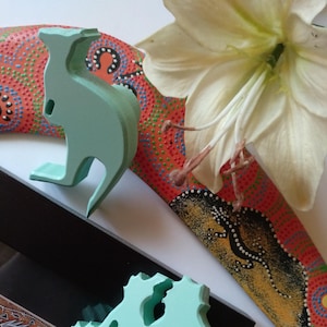 Puede incluir: Un conjunto de dos figuritas de madera con temática australiana. Las figuritas están pintadas de color verde claro. Una figurita es un canguro y la otra es un mapa de Australia. Las figuritas están sentadas sobre una superficie negra con un bumerán estampado en rojo, amarillo y negro en el fondo.
