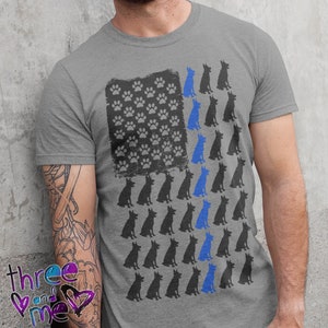 May include: A grey t-shirt with a blue line American flag design featuring paw prints and silhouettes of dogs. The flag has a thin blue line down the middle.