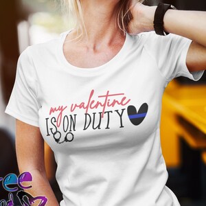 May include: White t-shirt with the text "my valentine is on duty" in pink and black lettering. A blue line heart is to the right of the text. Handcuffs are below the text.