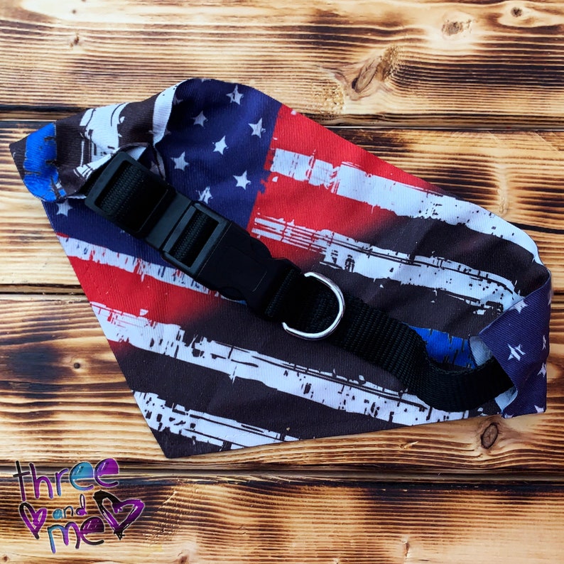 May include: A black and blue distressed American flag bandana with a black adjustable collar and silver ring. The bandana is on a wooden surface.