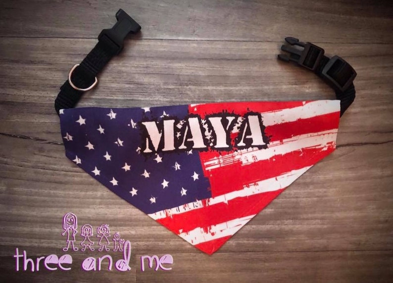 May include: A red, white, and blue bandana with a distressed American flag design. The bandana has the name "MAYA" printed in white on it. The bandana is attached to a black dog collar.