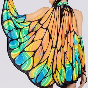 Puede incluir: Una colorida capa de alas de mariposa con cuello halter. La capa presenta un diseño vibrante con tonos naranja, amarillo, verde y azul, delineados en negro. Las alas están hechas de una tela transparente y ligera.