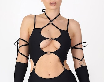 May include: Black bodysuit with cut-out details and silver ring accents. The halter top features a series of circular cutouts and straps. Long sleeves with finger holes and arm straps complete the look. The model is wearing orange-tinted sunglasses.