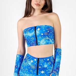 Può includere: Un top corto e pantaloncini coordinati blu e bianchi con una stampa celeste con stelle, lune e simboli zodiacali. Il top e i pantaloncini hanno cerniere nere sul davanti.