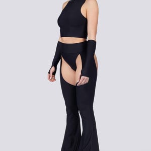 May include: Black two-piece outfit featuring a sleeveless crop top with a high collar and a zipper. The outfit includes matching arm sleeves and flared pants with open sides. The model is wearing black platform boots.