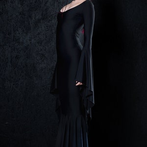 May include: A black long-sleeved dress with a fitted bodice and a flowing skirt. The dress has a scoop neckline and bell sleeves. The model is wearing the dress with black platform shoes.