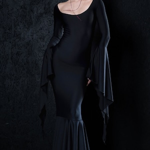 May include: A black long-sleeved dress with a fitted bodice and a flowing skirt. The dress has a scoop neckline and long, flowing sleeves. The model is wearing the dress with a simple necklace.
