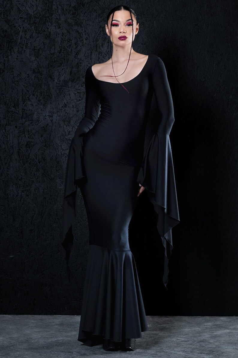 May include: A black long-sleeved dress with a fitted bodice and a flowing skirt. The dress has a scoop neckline and long, flowing sleeves. The model is wearing the dress with a simple necklace.