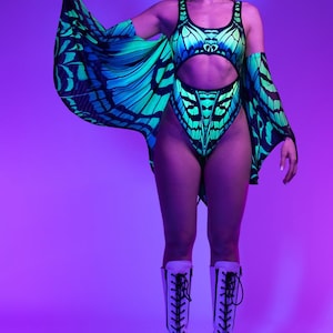 May include: A neon green and blue butterfly-themed bodysuit with matching cape sleeves and a choker. The outfit is paired with white platform boots. The design features a butterfly wing pattern. The background is a gradient of purple and pink.