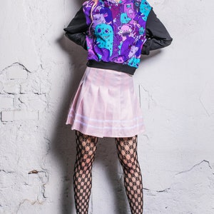 以下が含まれることがあります： ピンクのスカート、黒いフィッシュネットタイツ、黒いブーツを履いた女性。彼女は、漫画のモンスターのグラフィックプリントが施された紫と黒のパーカーを着ています。彼女はピンクの髪をしていて、花冠をかぶっています。