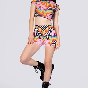 May include: A two-piece outfit featuring a crop top and high-waisted shorts. The set is pink with a colorful, wavy pattern and small hearts. The top has short sleeves and a high neckline. The shorts have a similar design. The model is wearing black platform boots.