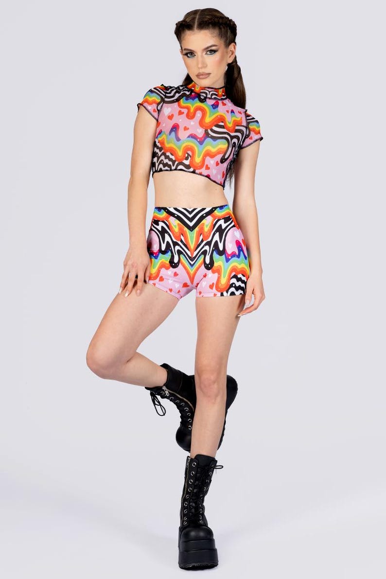 May include: A two-piece outfit featuring a crop top and high-waisted shorts. The set is pink with a colorful, wavy pattern and small hearts. The top has short sleeves and a high neckline. The shorts have a similar design. The model is wearing black platform boots.