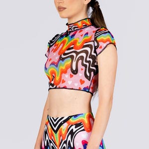 May include: A colorful two-piece outfit featuring a crop top and shorts. The top has a high neck and short sleeves, with a rainbow and black wavy pattern over a pink background with small red hearts. The shorts have a matching design.