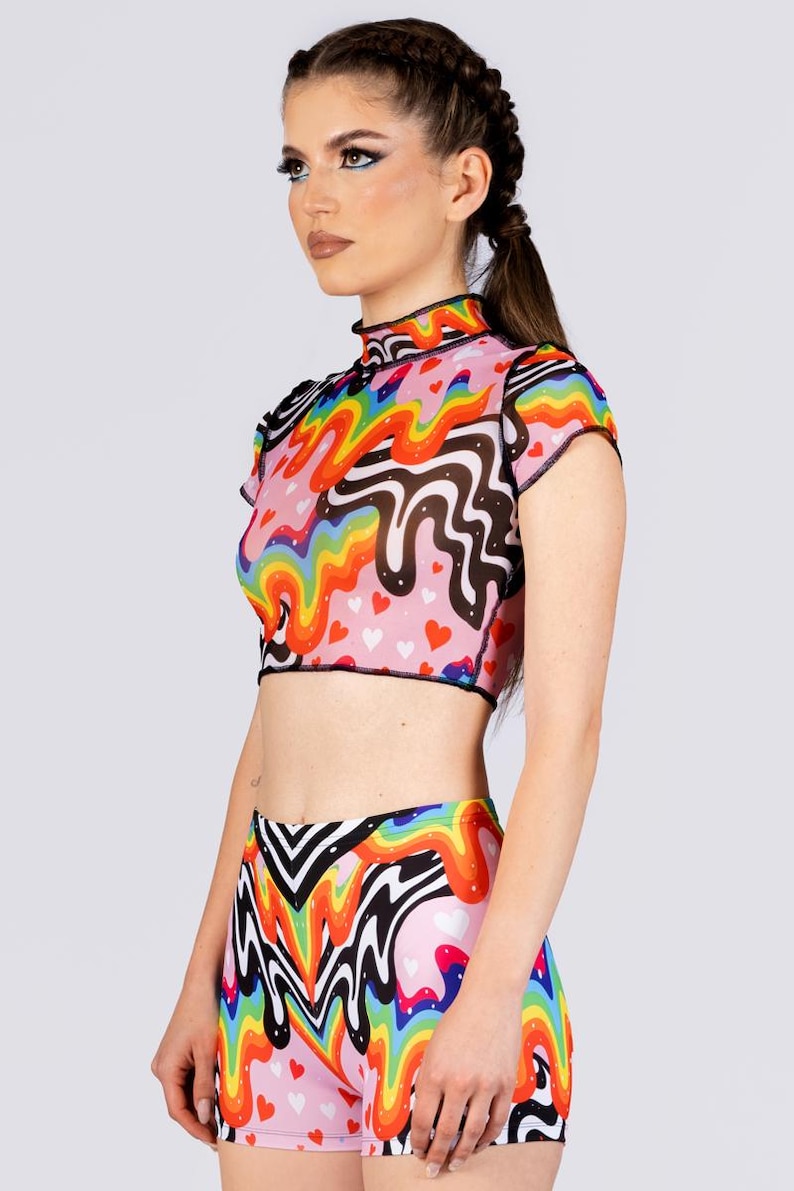 May include: A colorful two-piece outfit featuring a crop top and shorts. The top has a high neck and short sleeves, with a rainbow and black wavy pattern over a pink background with small red hearts. The shorts have a matching design.