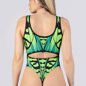 May include: A one-piece swimsuit featuring a butterfly wing design in shades of green and black. The swimsuit has a scoop neckline, a cut-out at the waist, and a thong back. The model is facing away from the camera.