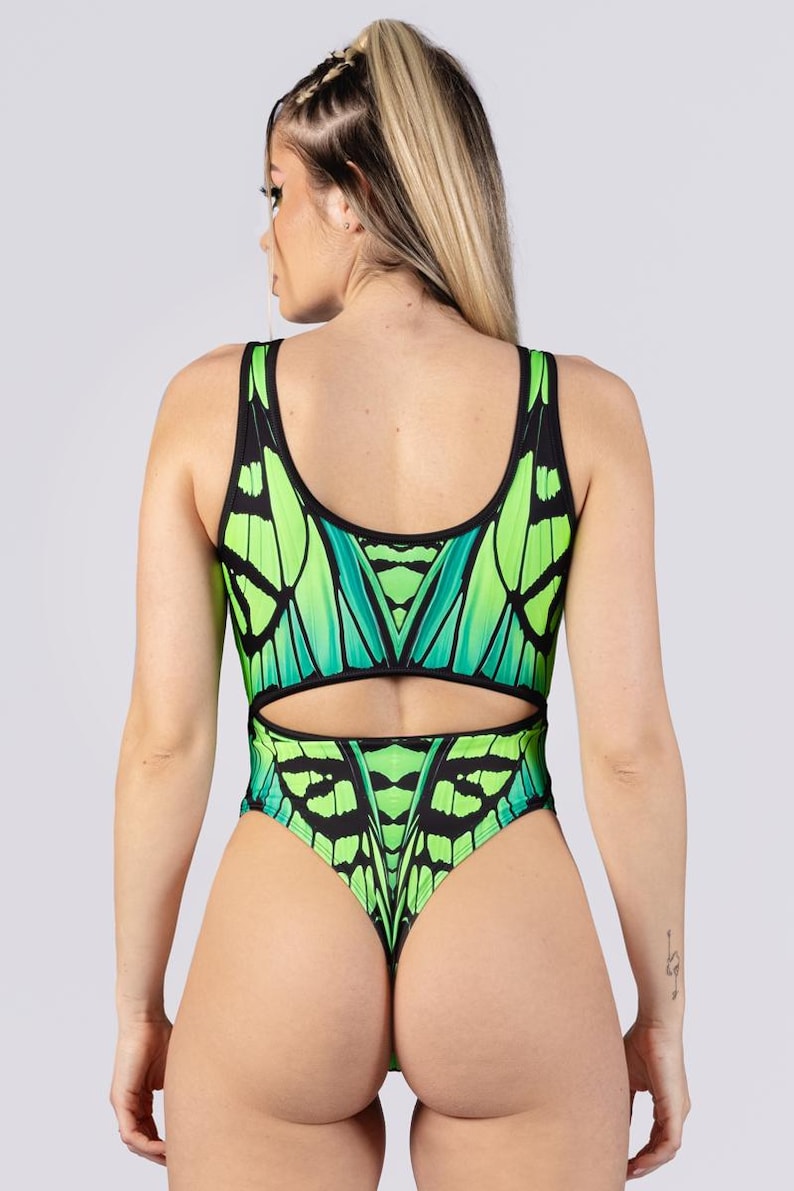May include: A one-piece swimsuit featuring a butterfly wing design in shades of green and black. The swimsuit has a scoop neckline, a cut-out at the waist, and a thong back. The model is facing away from the camera.