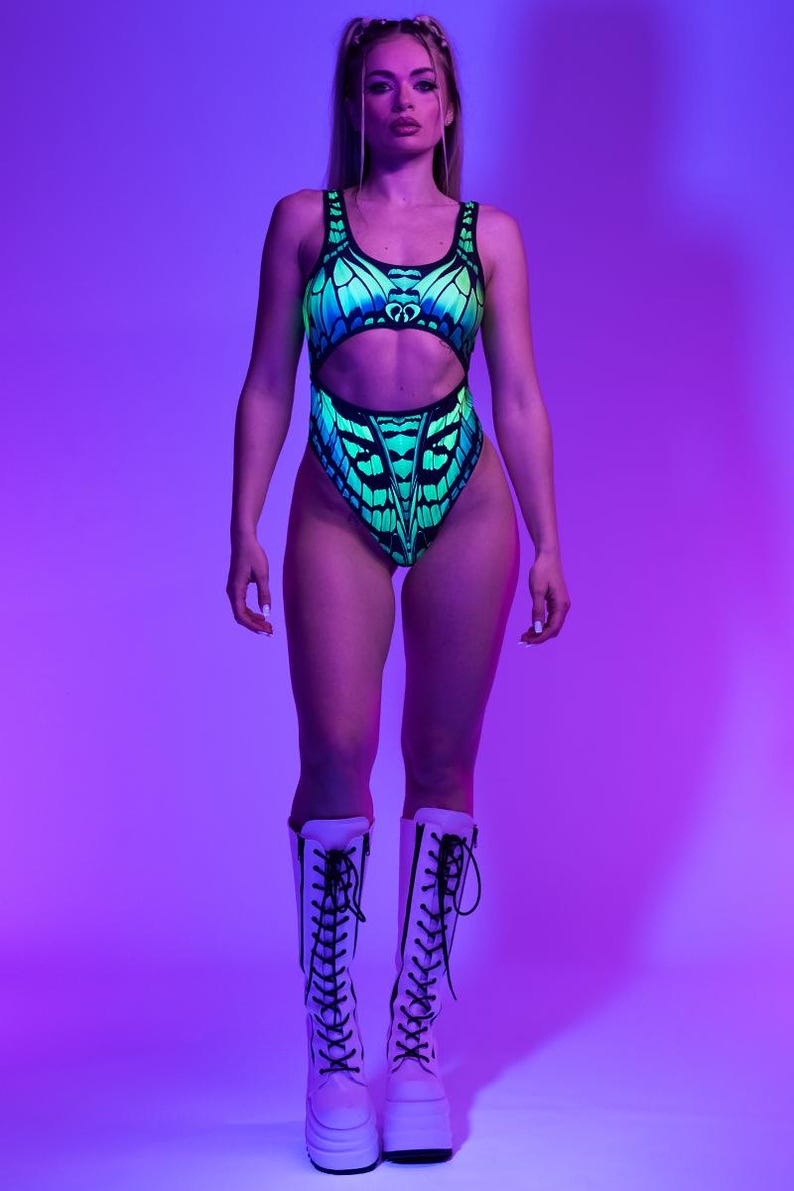 May include: A one-piece swimsuit with a butterfly wing design in shades of blue and green. The swimsuit has a cut-out design at the waist and is paired with white platform boots with black laces. The background is a gradient of purple and blue.