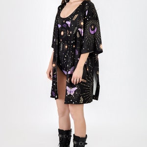 May include: A black kimono with a celestial print featuring butterflies, moons, stars, and suns. The kimono has long sleeves and a side slit. The model is wearing black boots.