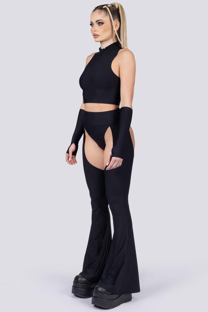 May include: Black two-piece outfit featuring a sleeveless crop top with a high collar and a zipper. The outfit includes matching arm sleeves and flared pants with open sides. The model is wearing black platform boots.