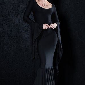 May include: A woman wearing a black long-sleeved dress with a fitted bodice and a flowing skirt. The dress has a scoop neckline and long bell sleeves. The woman is standing in front of a black textured background.