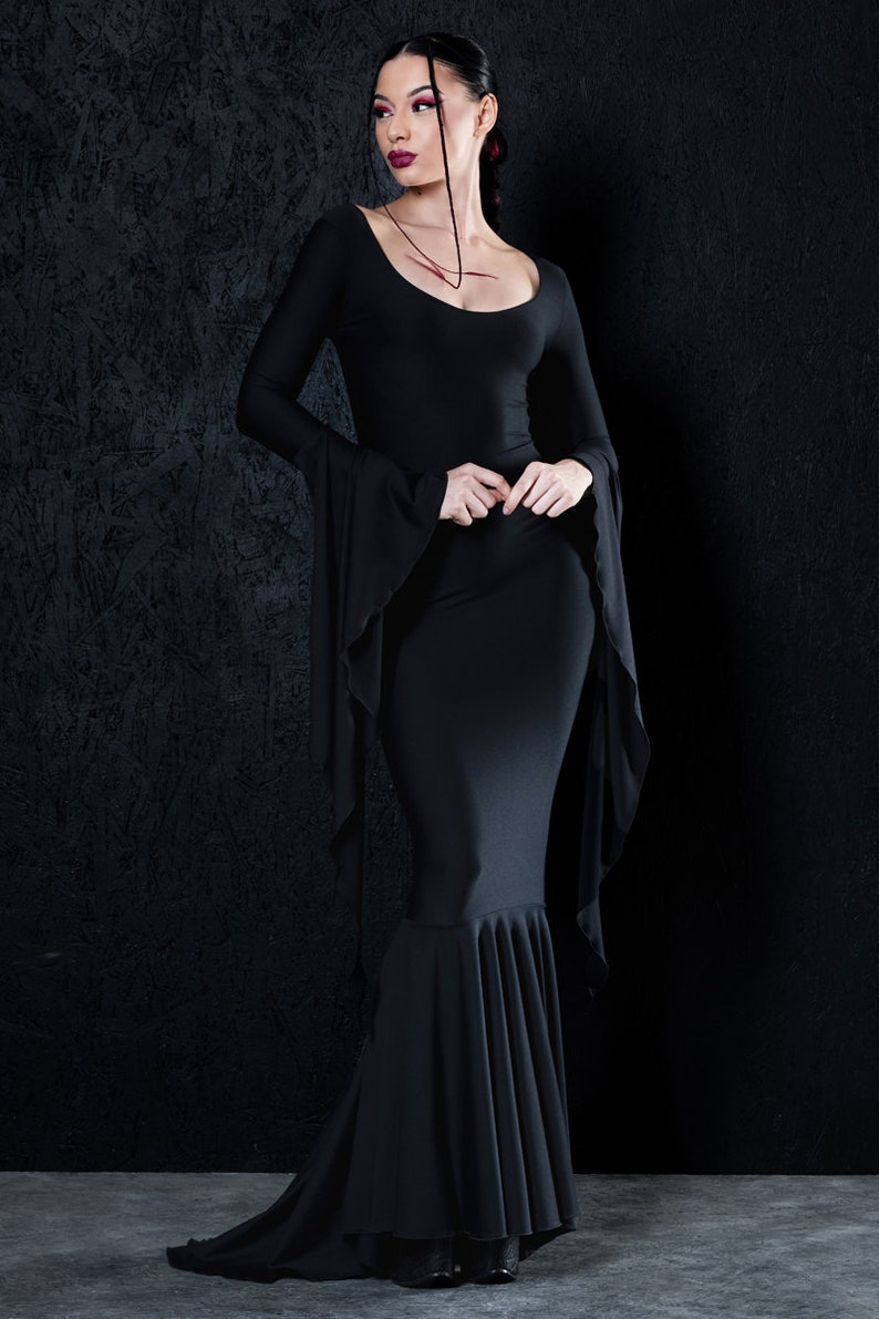 May include: A woman wearing a black long-sleeved dress with a fitted bodice and a flowing skirt. The dress has a scoop neckline and long bell sleeves. The woman is standing in front of a black textured background.