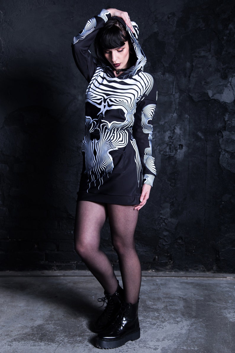 Puede incluir: Un vestido negro con capucha y estampado gr&aacute;fico de calavera blanca. El vestido tiene mangas largas y una silueta ajustada. La modelo lleva medias negras y botas de plataforma negras.