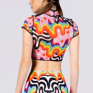May include: A pink crop top and matching shorts set with a colorful rainbow and heart pattern. The top has short sleeves and a high neck, with black trim. The shorts are high-waisted and feature the same design.