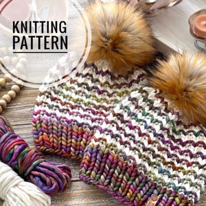 May include: Two knitted hats with colorful yarn and faux fur pom poms. The hats are white with a colorful chevron pattern. The yarn is a mix of purple, pink, green, and brown. The hats are on a wooden surface with a spool of yarn and a wooden bead garland.