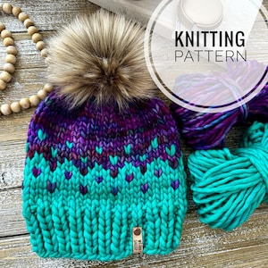 May include: A hand-knitted beanie in shades of teal, purple, and brown, topped with a fluffy faux fur pom-pom. The hat features a heart-shaped pattern and the text "KNITTING PATTERN". A wooden tag reads "WEIRD SHEEP".