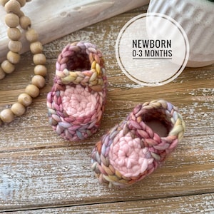 Può includere: Un paio di scarpine fatte a mano per neonati nei toni del rosa, giallo e viola. Le scarpine sono all'uncinetto con un filato strutturato e presentano un design rotondo sulla parte superiore. Il testo "NEWBORN 0-3 MONTHS" è in un cerchio bianco.