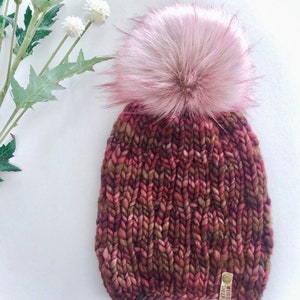 May include: A chunky knit beanie with a pink faux fur pom pom. The beanie is made of a thick, textured yarn in shades of brown, red, and pink. The beanie has a brown leather tag with the text "WOVEN SHEEP" on it.