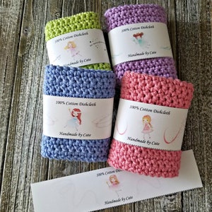 May include: Five crocheted dishcloths in different colors, including green, purple, blue, pink, and a white tag. Each dishcloth has a label that reads "100% Cotton Dishcloth" and "Handmade by Cate".