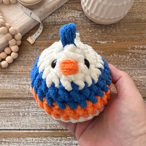 May include: A handmade, crocheted bird toy in white, blue, and orange. The bird has black safety eyes and a small orange beak. The toy is held in a hand, showcasing the soft texture of the yarn. A charming, whimsical design.