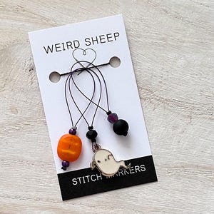 May include: Set of four stitch markers with Halloween themes. The markers feature a ghost, a pumpkin, a black bead, and a purple bead. The markers are attached to a white card with the text "Weird Sheep Stitch Markers".