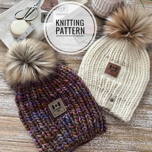 May include: Two knitted hats with faux fur pom poms. One hat is a multi-colored yarn with a brown leather patch that says "Elbows Up!" The other hat is white with a brown leather patch that says "Elbows Up!" Both hats are on a wooden surface.