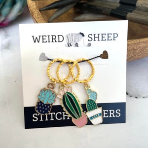 May include: A set of three gold stitch markers with cactus charms. The charms feature cacti and succulents, with blue, green, pink, and white enamel details. The card reads "WEIRD SHEEP" and "STITCHERS".