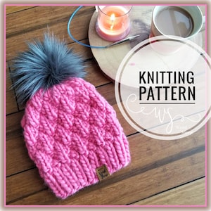 May include: Pink knitted hat with a gray pom pom. The hat has a textured stitch pattern and a brown leather tag with the words "Weird Sheep" on it. The image also includes a cup of coffee, a lit candle, and a knitting needle.