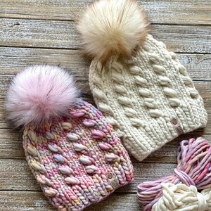 Puede incluir: Dos gorros de punto con pompones esponjosos. Un gorro es blanco con un patr&oacute;n de punto de cable y el otro es de hilo multicolor con un patr&oacute;n de punto grueso. Ambos gorros tienen una etiqueta de cuero con el texto "The Wool Sheep" en ellos.